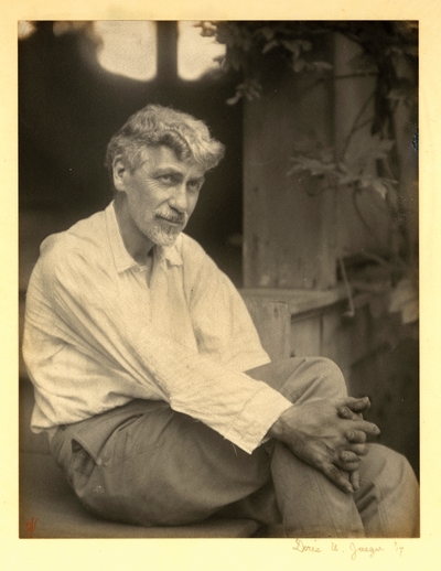 Charles R. Lamb;  Bearded man with mustache, in shirt and pants, seated with hands clasped around knee, with plant in background