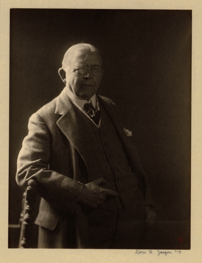 Elderly man in glasses, tie, and suit with handkerchief, standing with elbow leaning on back of chair