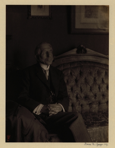 Elderly man with sideburns, in suit and tie with glasses hanging from chain, seated on sofa with pictures hanging behind it