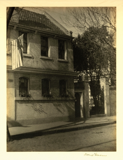 Brick and stone building with tiled roof, planters on windowsills, and iron gate, flying American flag