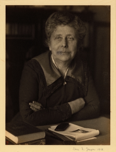 Woman seated at desk with arms crossed and books, paper, and other objects on desk