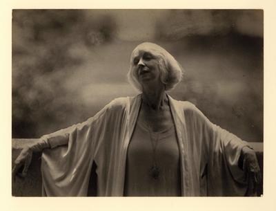 Ruth St. Denis; Woman in loose robe with large pendant, leaning against low wall, with head tilted back