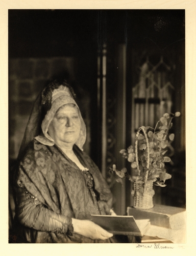 Woman in lace shawl and mantilla, seated beside plant in basket, holding book.  Ca. 1918-25