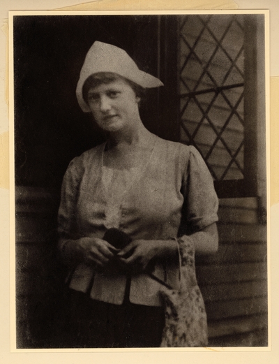 Young woman in Dutch costume, standing in front of open window, with bag hanging on her arm