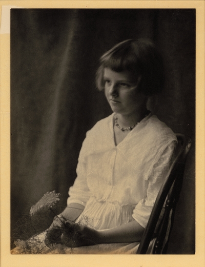 Girl in necklace and dress, seated in chair, holding flower