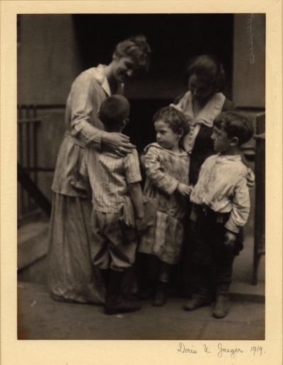 Three children, one carrying books, standing with two women (teachers?)