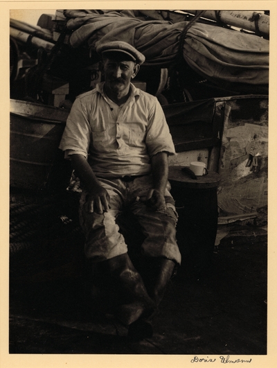 Man with mustache, in hat, ragged pants, and boots, seated with mug on a barrel beside him