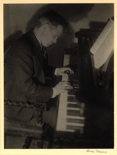 John Jacob Niles, in suit and tie, playing piano, looking down.  1932-1933