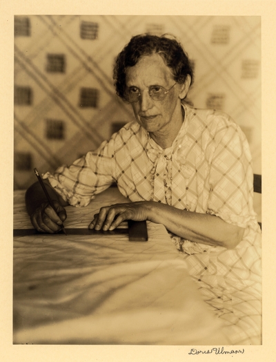 Mrs. C. D. Hood [or Wood], Dalton La.  Elderly woman in glasses and dress, seated at table with pencil and T-square, with quilt in background