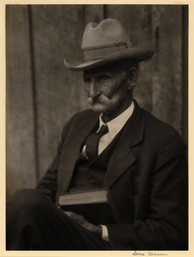 Elderly man with mustache, in hat, suit, and tie, seated with book