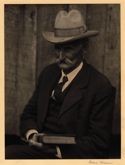 Elderly man with mustache, in hat, suit, and tie,  seated in chair with book, looking down