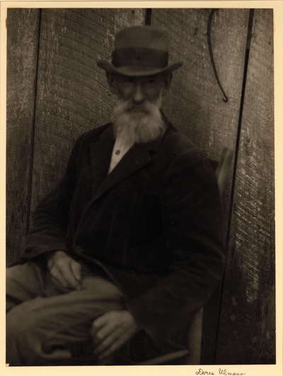 Elderly, bearded man in hat and coat, seated in chair