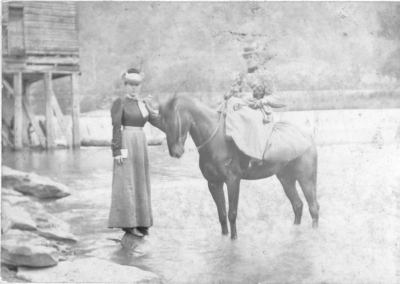 Two unidentified women, one is on a horseback.  Both are standing in a creek near waterfall.  The horse is laden with supplies. Bromide